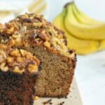 A loaf of banana nut bread with chopped walnuts on top sits on a wooden cutting board, with a bunch of bananas in the background.