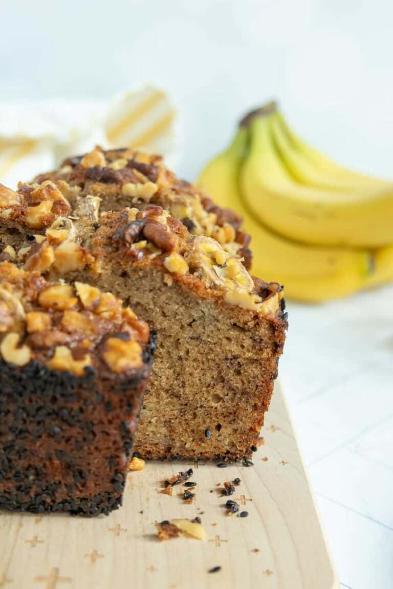 A loaf of banana nut bread with chopped walnuts on top sits on a wooden cutting board, with a bunch of bananas in the background.