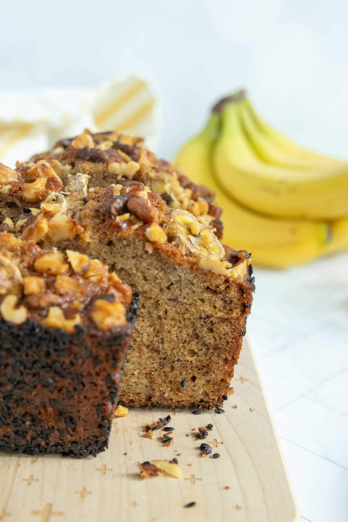 A loaf of banana nut bread with chopped walnuts on top sits on a wooden cutting board, with a bunch of bananas in the background.