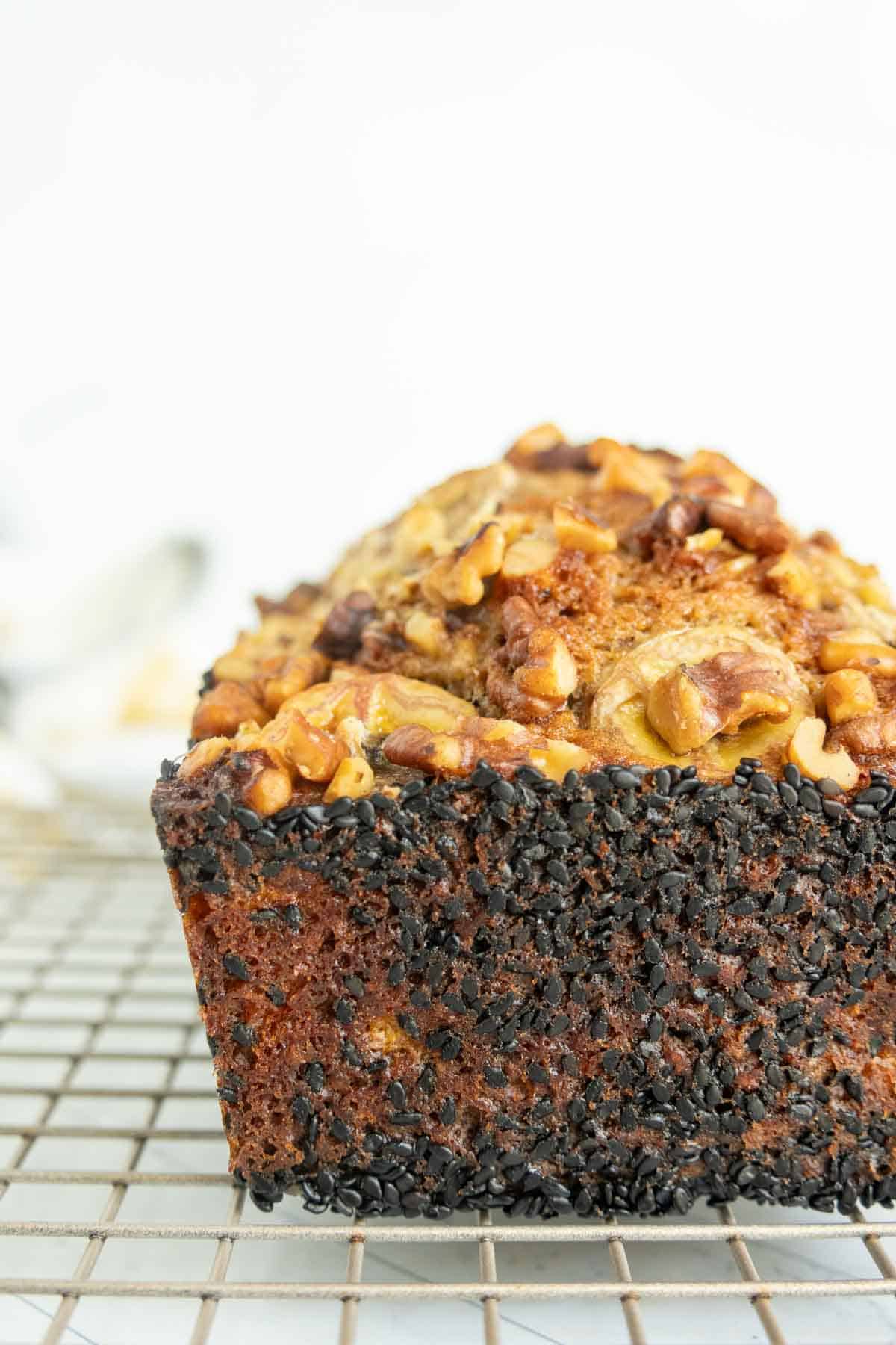 Close-up of a banana nut bread loaf topped with chopped nuts and coated with black sesame seeds along the sides, resting on a wire cooling rack.