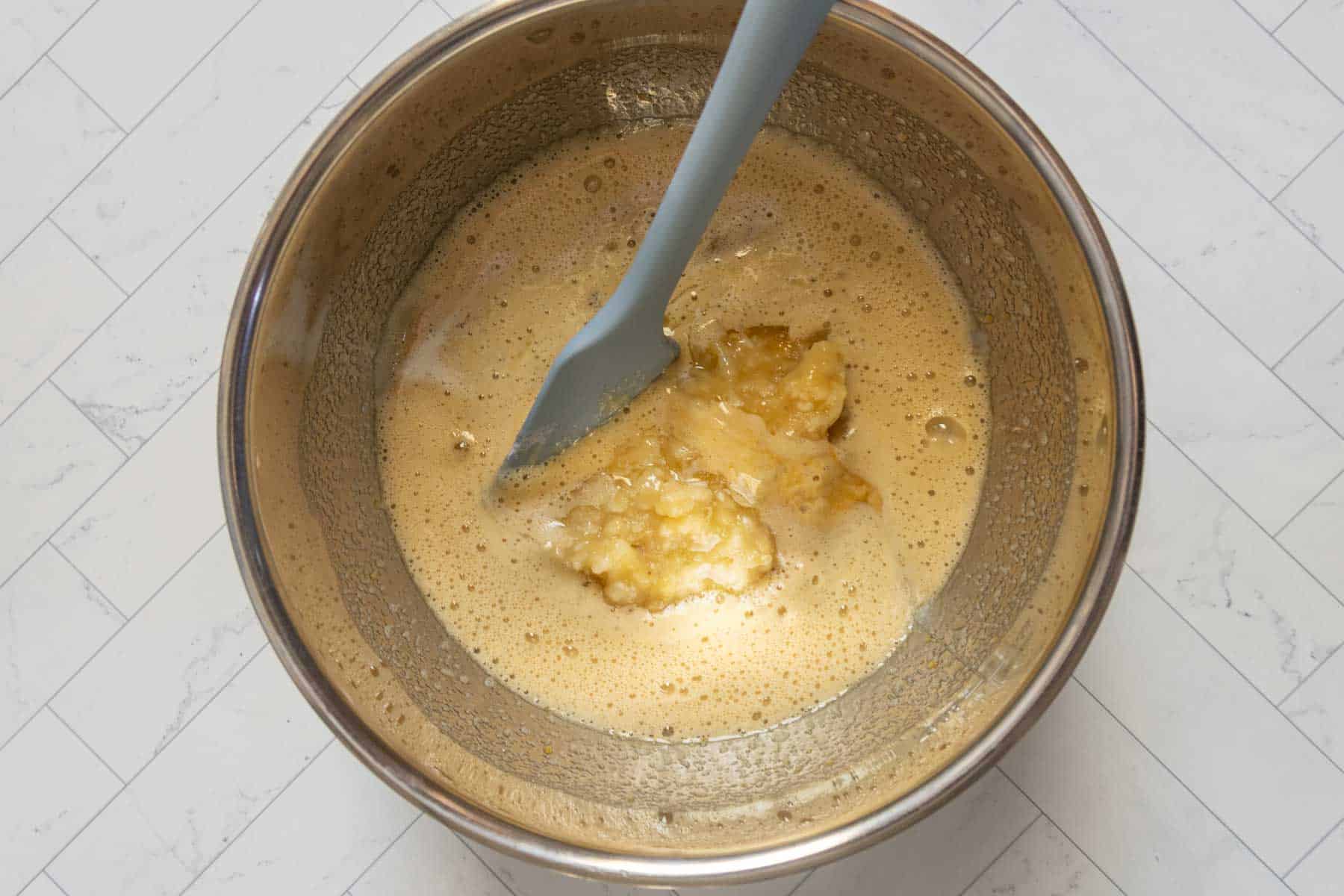 A metal mixing bowl containing a light brown banana nut bread mixture with mashed bananas, stirred with a gray spatula, on a white tiled surface.