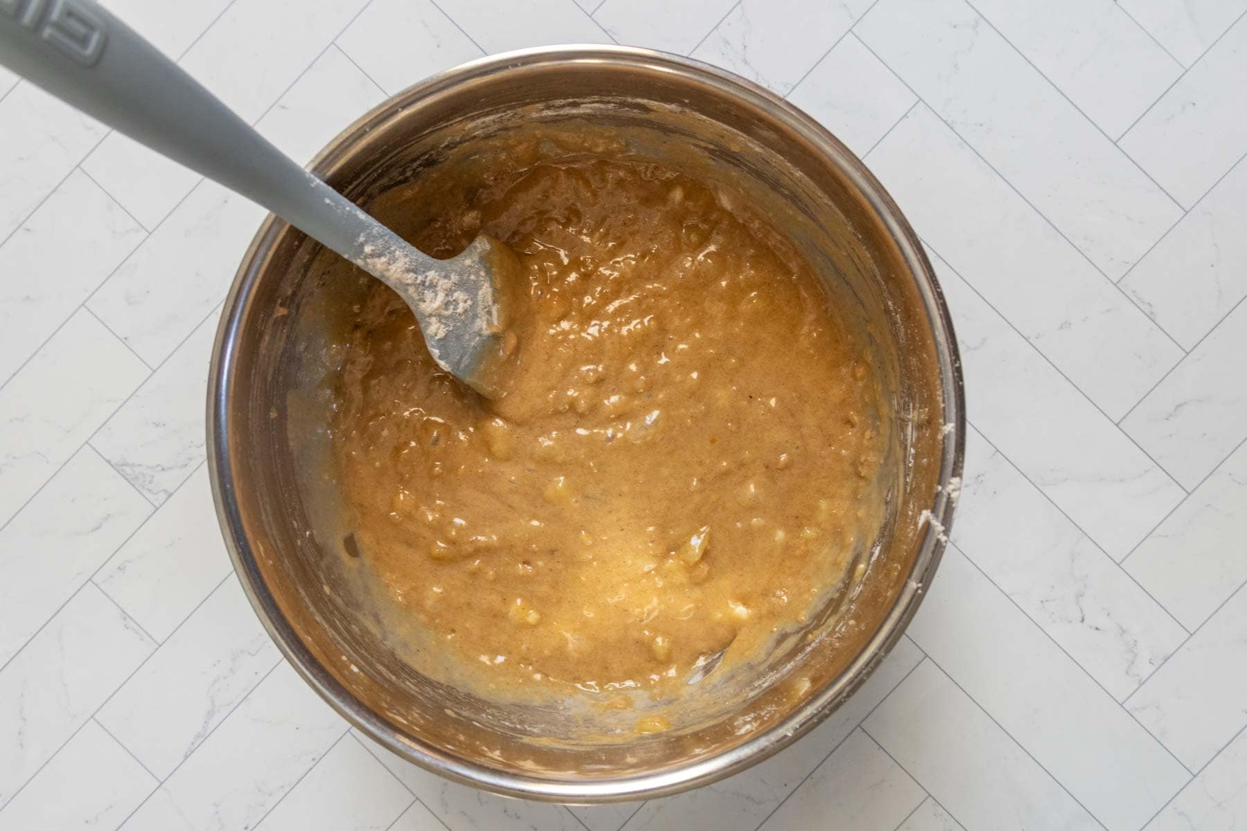 A metal bowl containing thick, light brown banana nut bread batter being mixed with a gray spatula, placed on a white tiled surface.