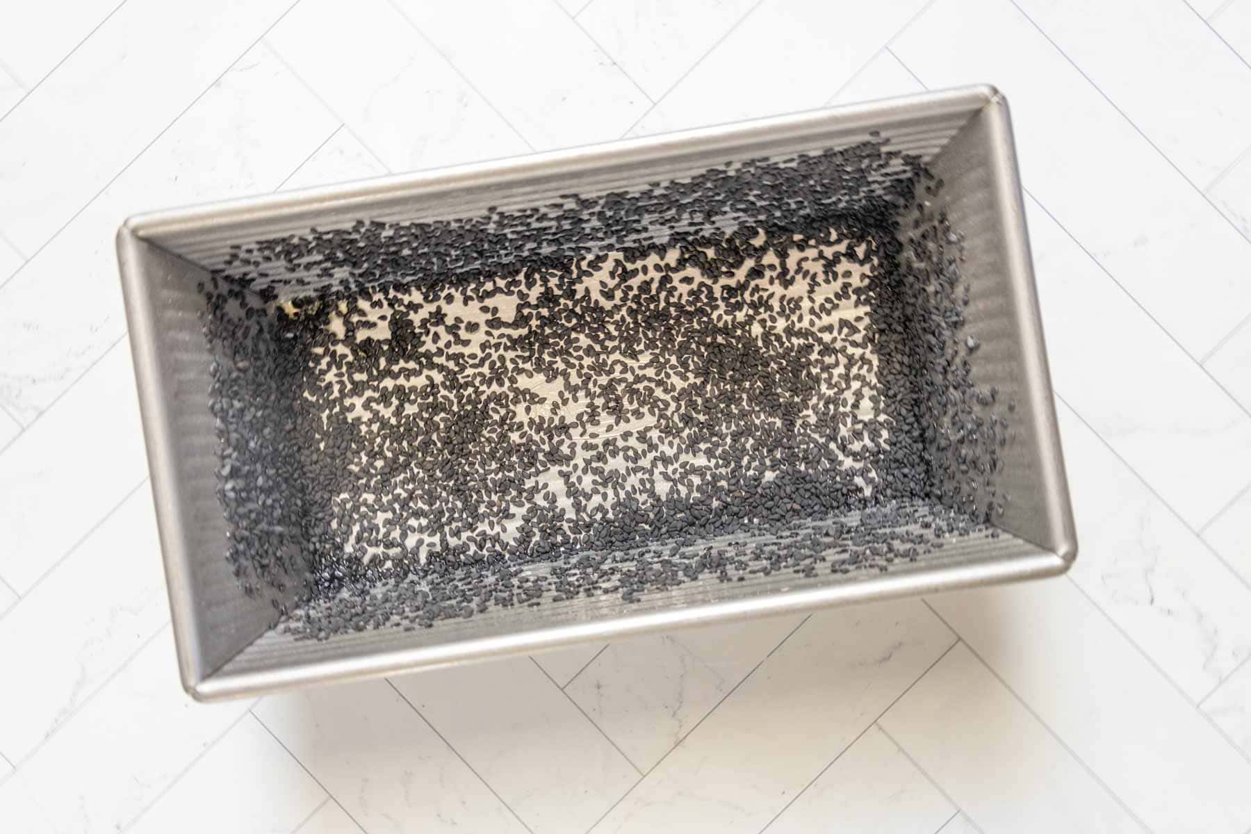 Rectangular metal baking pan with black poppy seeds scattered along the bottom and sides, ready for banana nut bread, placed on a white tiled surface.