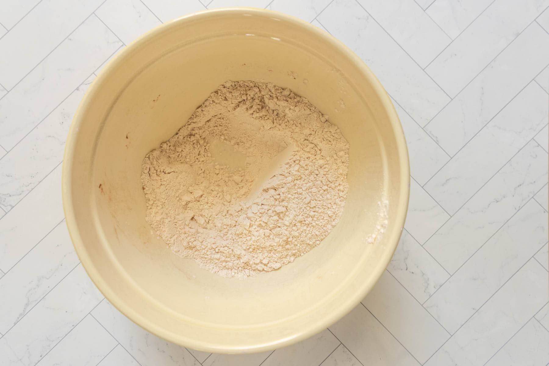 A large beige mixing bowl containing a mound of flour mixture for banana nut bread sits on a white, herringbone-patterned surface.
