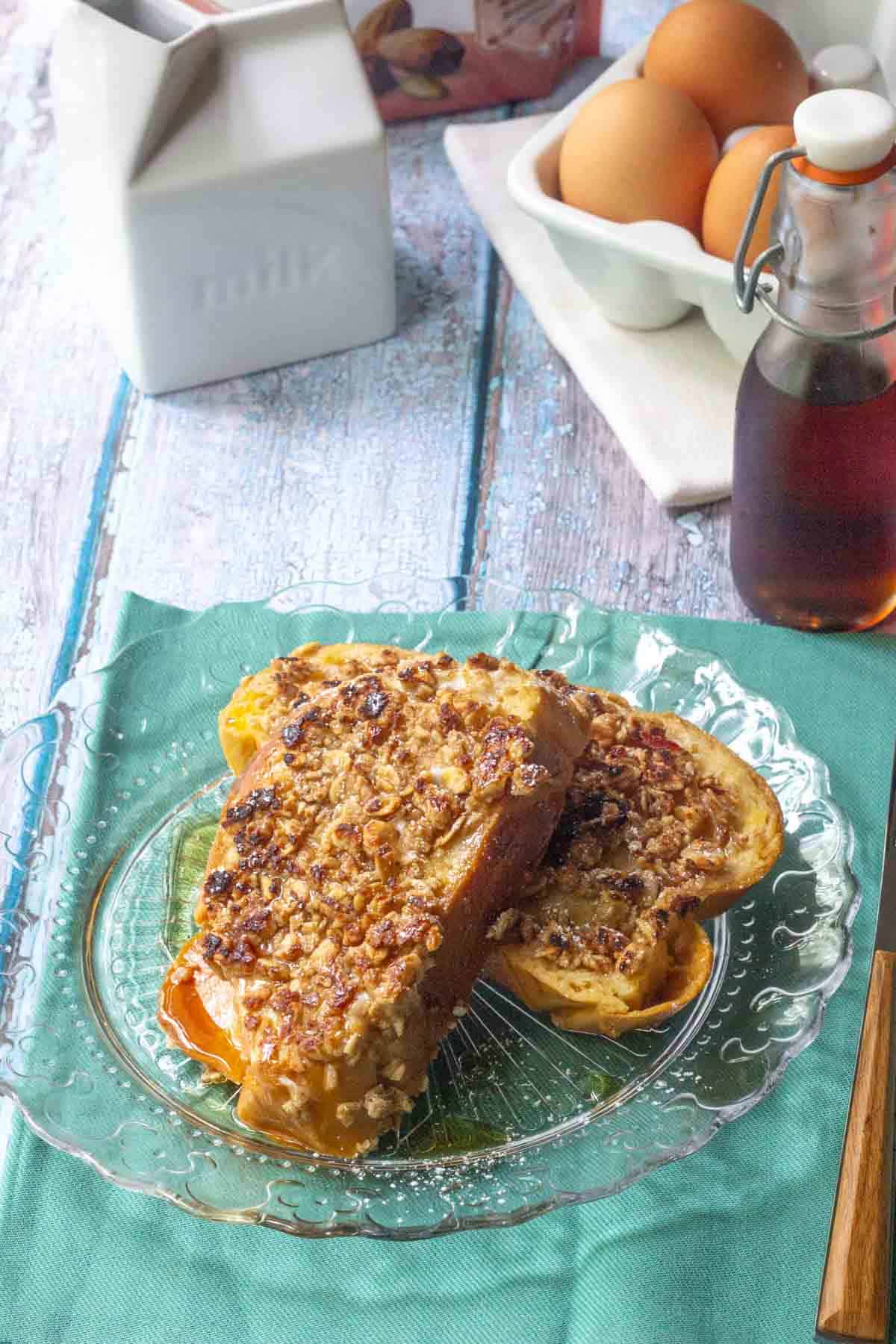 Two slices of granola French toast with a crumb topping are served on a glass plate, next to a bottle of syrup, a carton of eggs, and a white milk container.