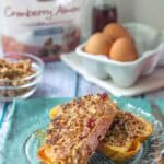 Two slices of granola French toast topped with crunchy granola on a glass plate, with eggs, syrup, and a "Cranberry Almond" bag featured in the background.