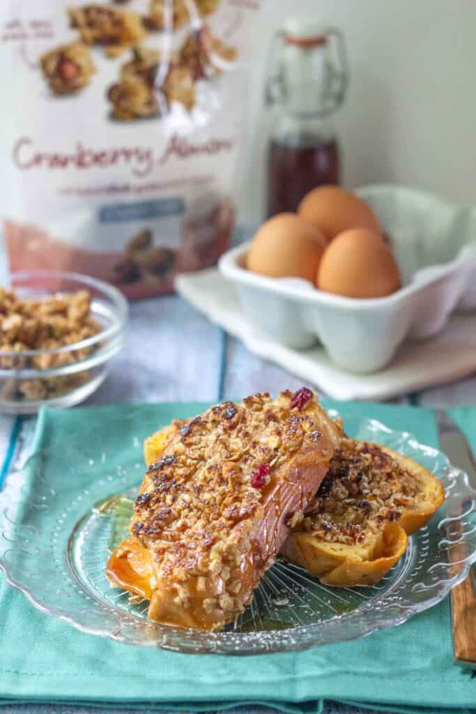 Two slices of granola French toast topped with crunchy granola on a glass plate, with eggs, syrup, and a "Cranberry Almond" bag featured in the background.