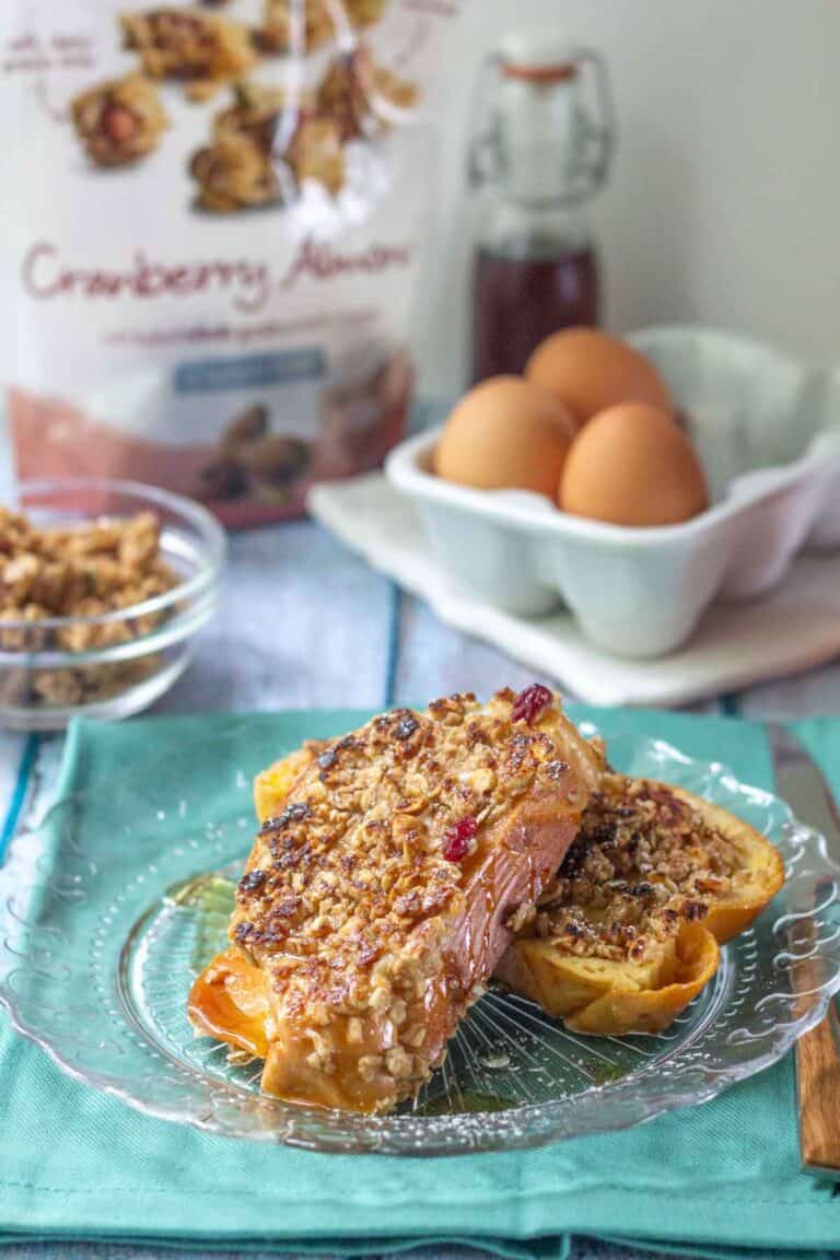 Two slices of granola French toast topped with crunchy granola on a glass plate, with eggs, syrup, and a "Cranberry Almond" bag featured in the background.