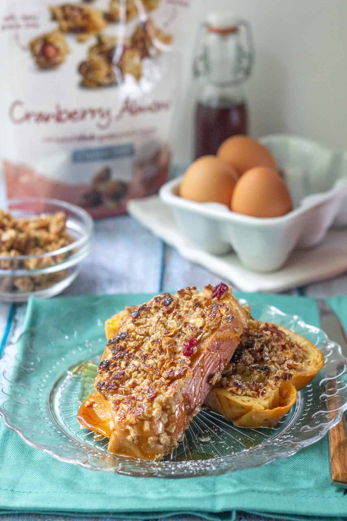 Two slices of granola French toast topped with crunchy granola on a glass plate, with eggs, syrup, and a "Cranberry Almond" bag featured in the background.