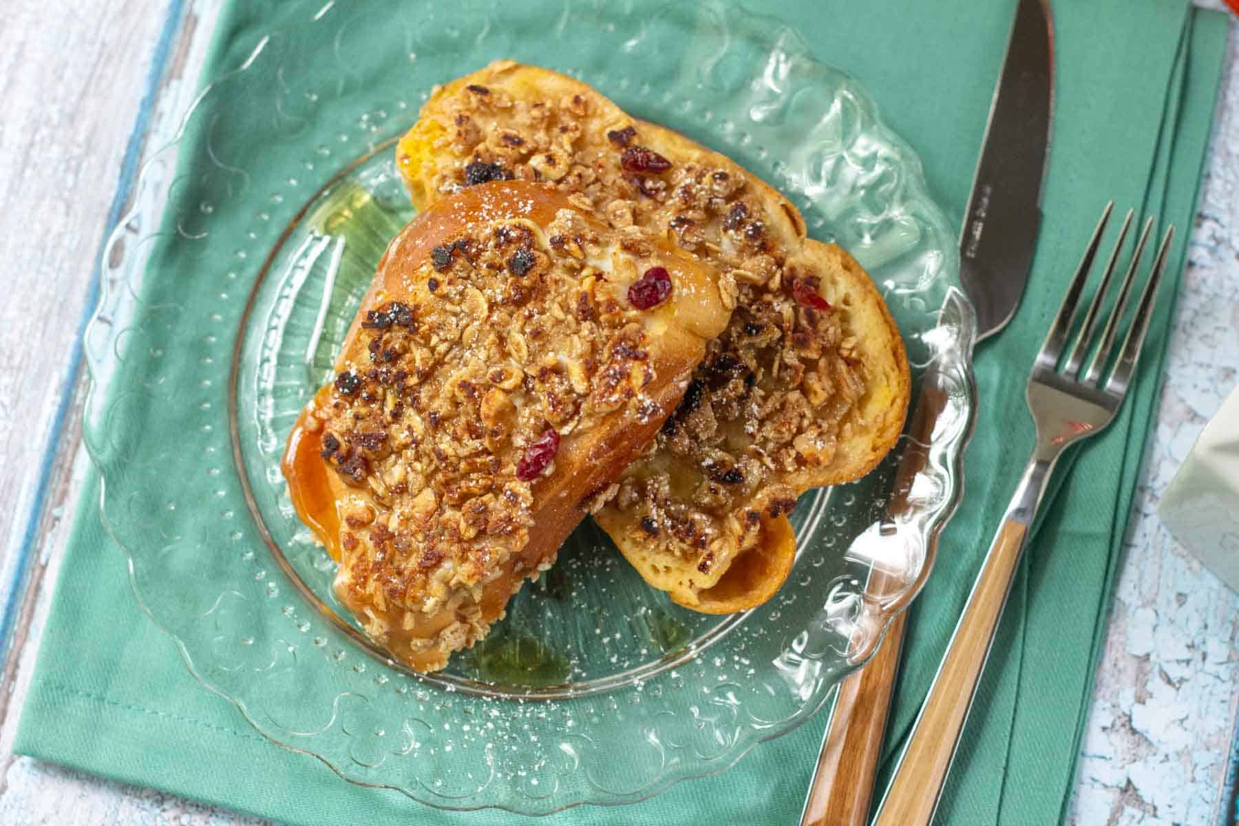 Two slices of granola French toast topped with dried fruit sit on a clear glass plate, set atop a green napkin with a knife and fork placed neatly beside it.