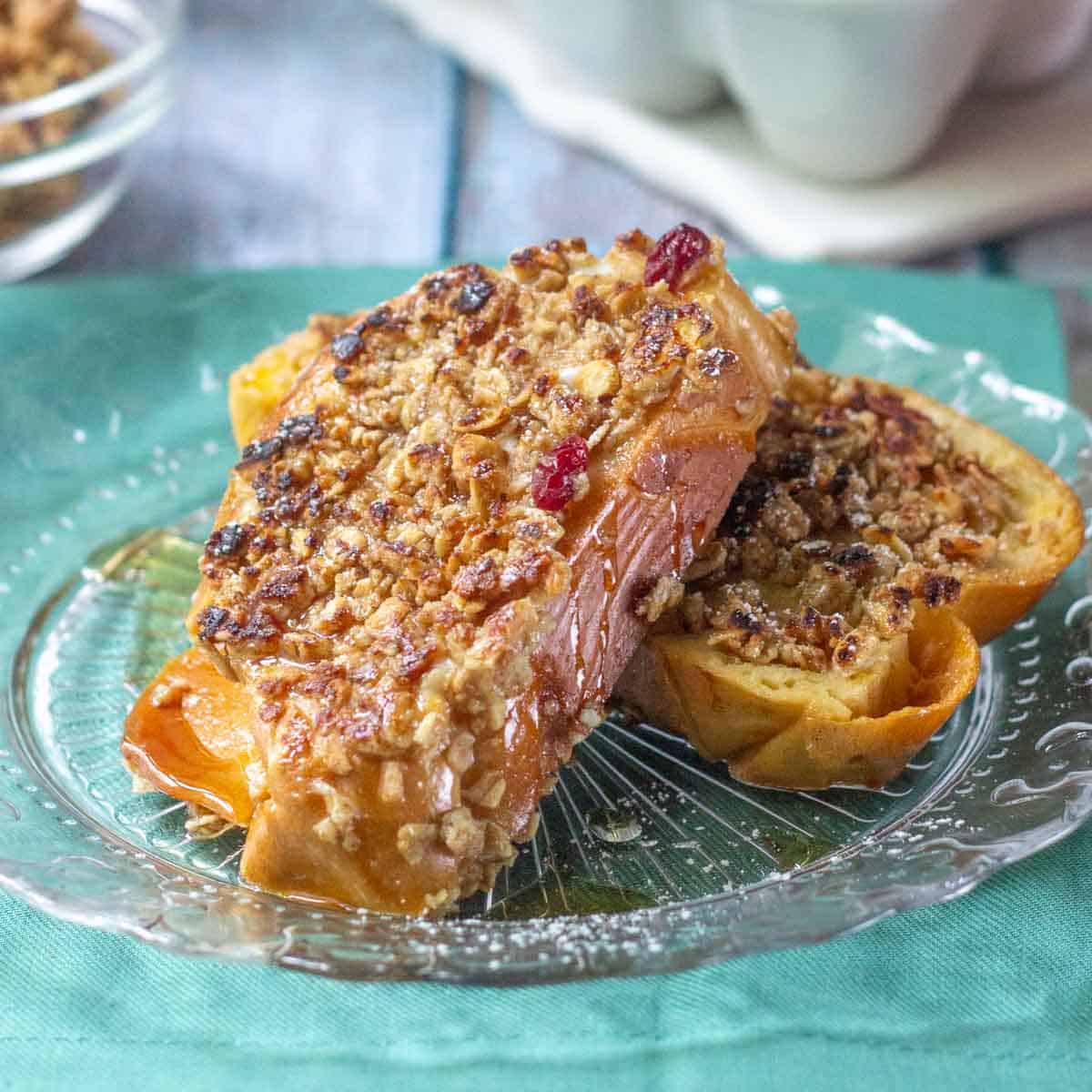 Two slices of baked French toast topped with a nutty, crumbly streusel sit on a clear glass plate atop a teal napkin—perfect inspiration for your favorite breakfast recipes.