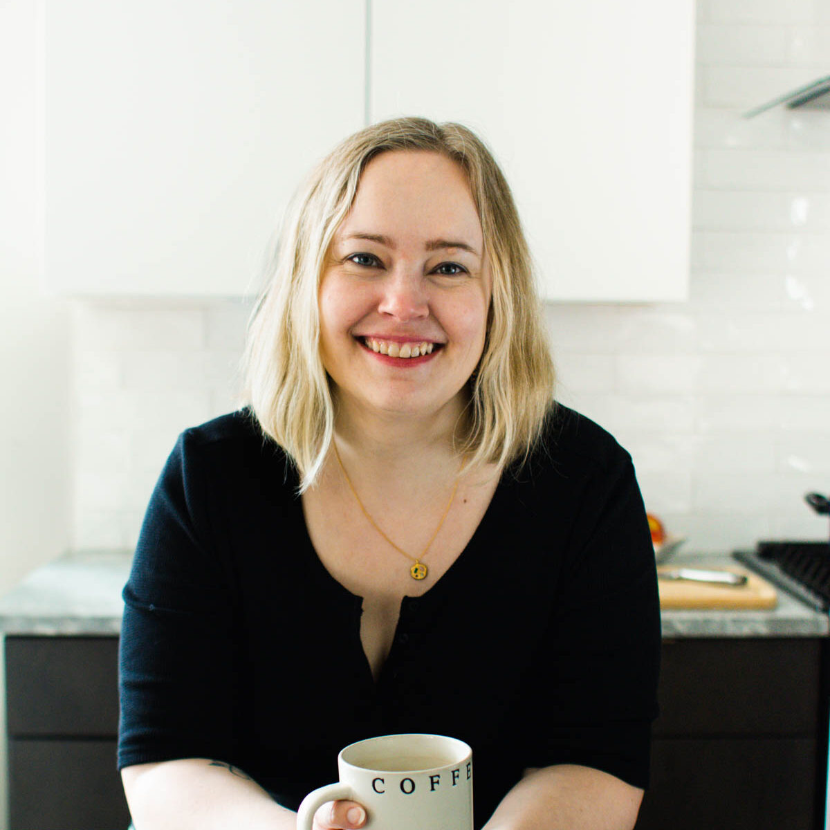 Woman with blonde hair in a black top smiles while holding a coffee mug in a modern kitchen with white cabinets.