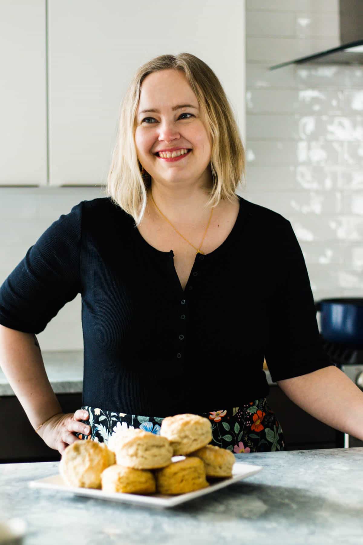 A woman with blonde hair stands in a kitchen, smiling, with a plate of biscuits on the counter in front of her—ready to share about simply breakfast recipes.