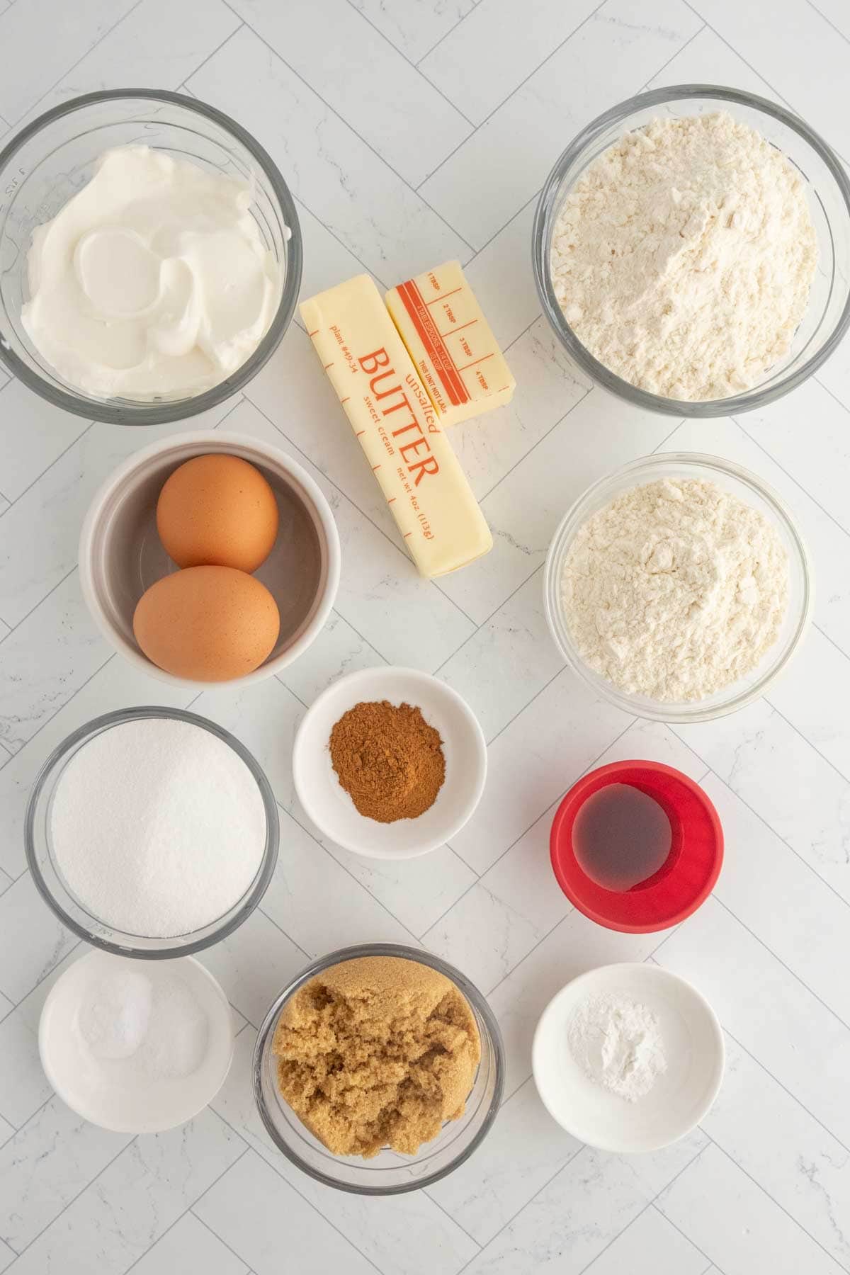Top-down view of baking ingredients for coffee cake muffins, including eggs, butter, flour, sugar, brown sugar, sour cream, spices, vanilla extract, and baking powder on a white tiled surface.
