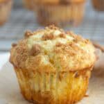 A close-up of a crumb-topped coffee cake muffin on a white plate, with more muffins blurred in the background on a cooling rack.