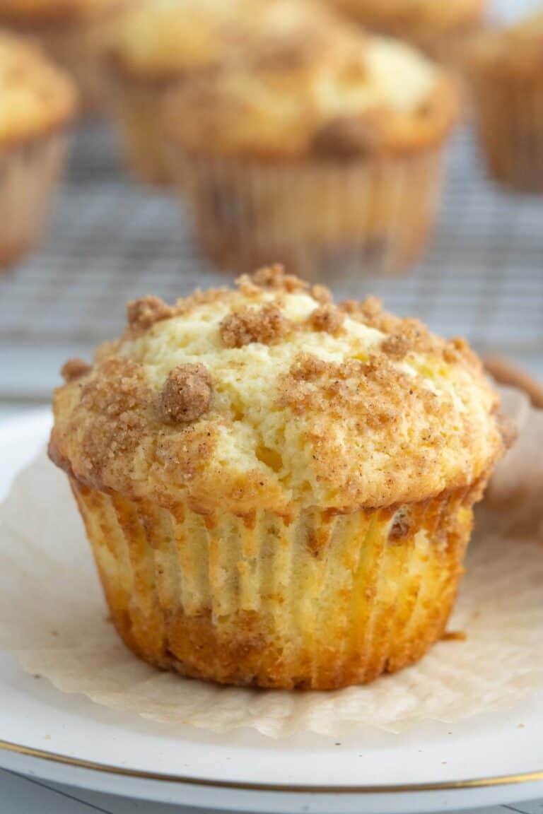 A close-up of a crumb-topped coffee cake muffin on a white plate, with more muffins blurred in the background on a cooling rack.