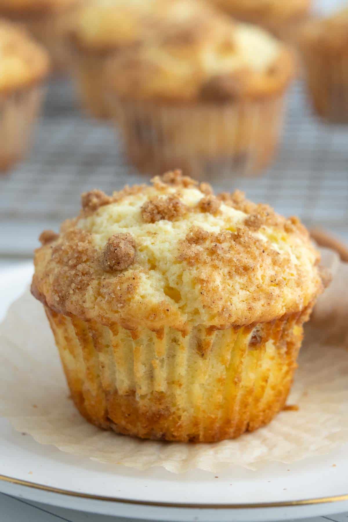A close-up of a crumb-topped coffee cake muffin on a white plate, with more muffins blurred in the background on a cooling rack.