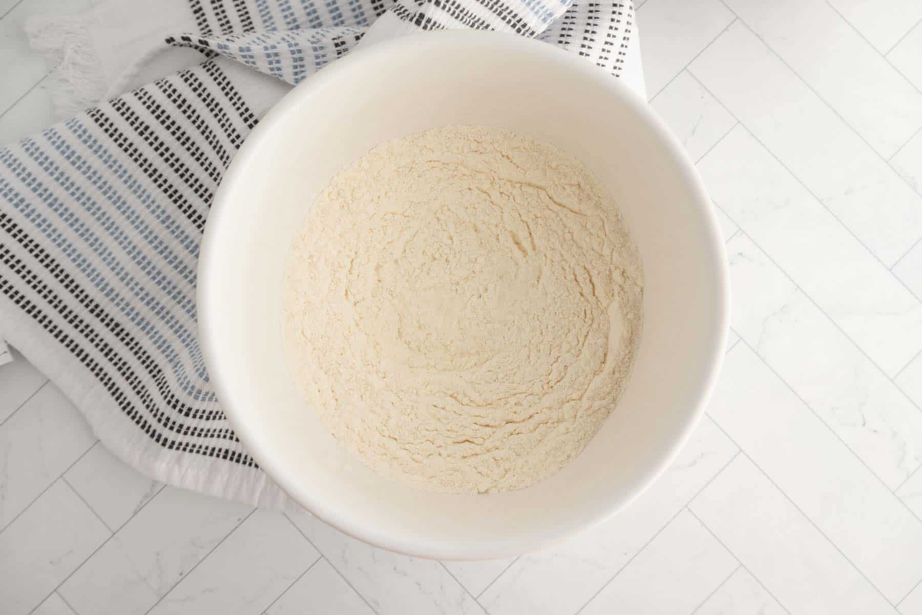 A white mixing bowl filled with dry flour mixture sits on a white tile countertop next to a striped kitchen towel, ready for baking delicious coffee cake muffins.