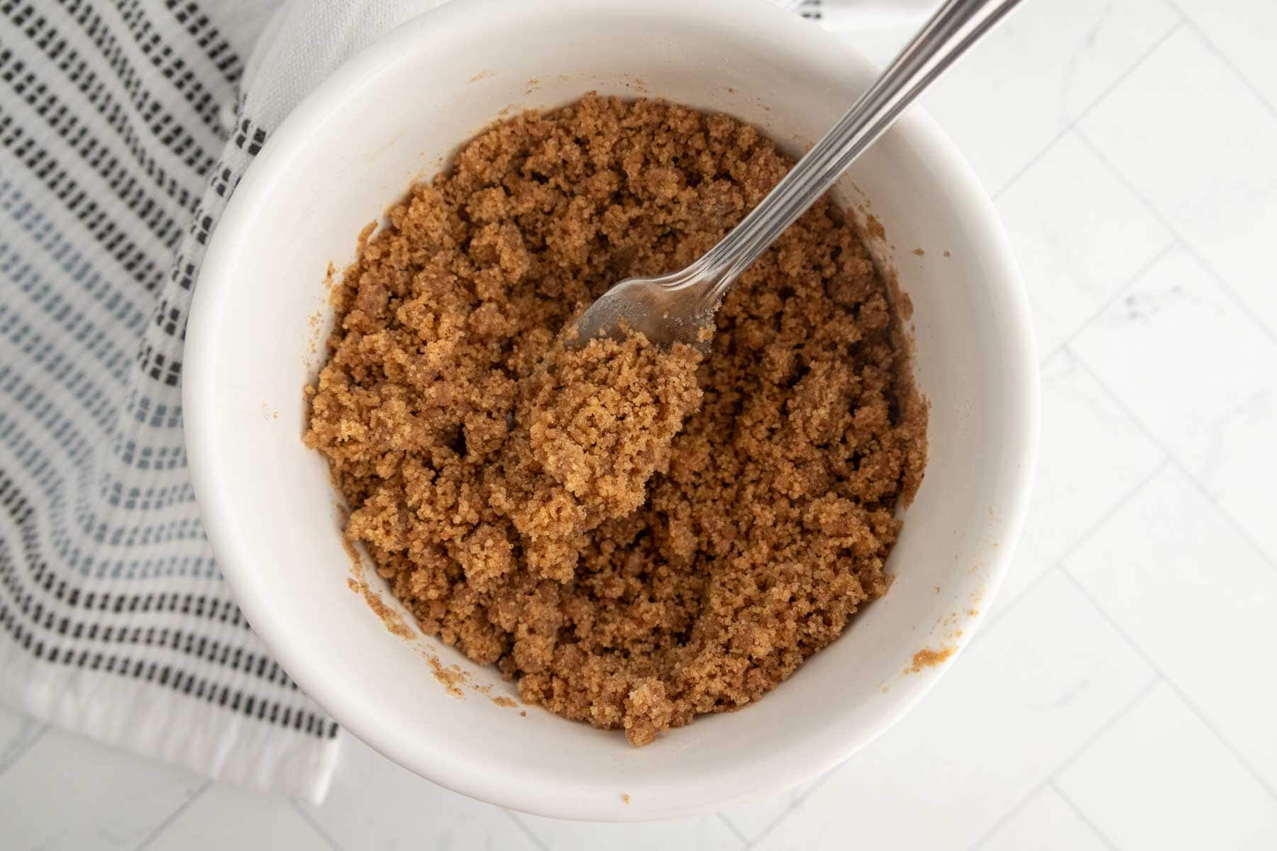 A white bowl filled with a crumbly brown sugar mixture for coffee cake muffins, with a fork resting inside, placed on a white countertop next to a striped cloth.