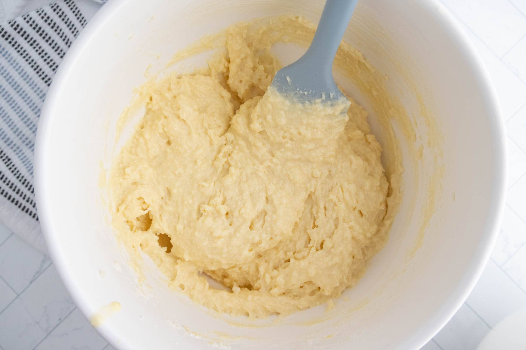 A white mixing bowl containing pale yellow coffee cake muffins batter being stirred with a light blue spatula, placed on a white countertop next to a striped towel.