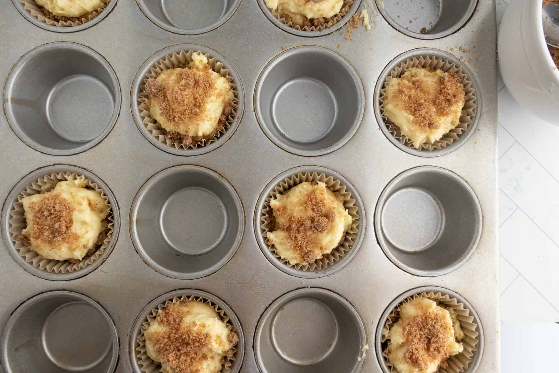 A muffin tin filled with paper liners, some holding unbaked coffee cake muffins topped with a brown sugar mixture, while other cups remain empty.