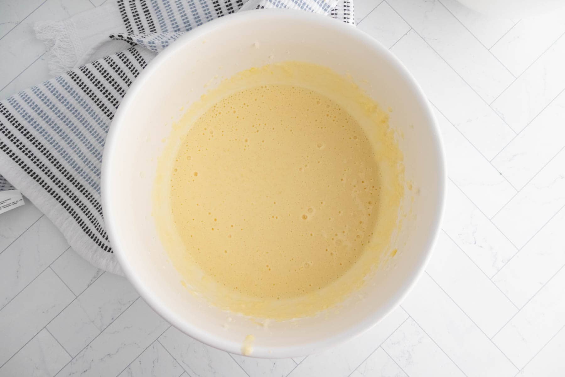 A white bowl filled with pale yellow coffee cake muffin batter sits on a white tiled surface next to a striped kitchen towel.