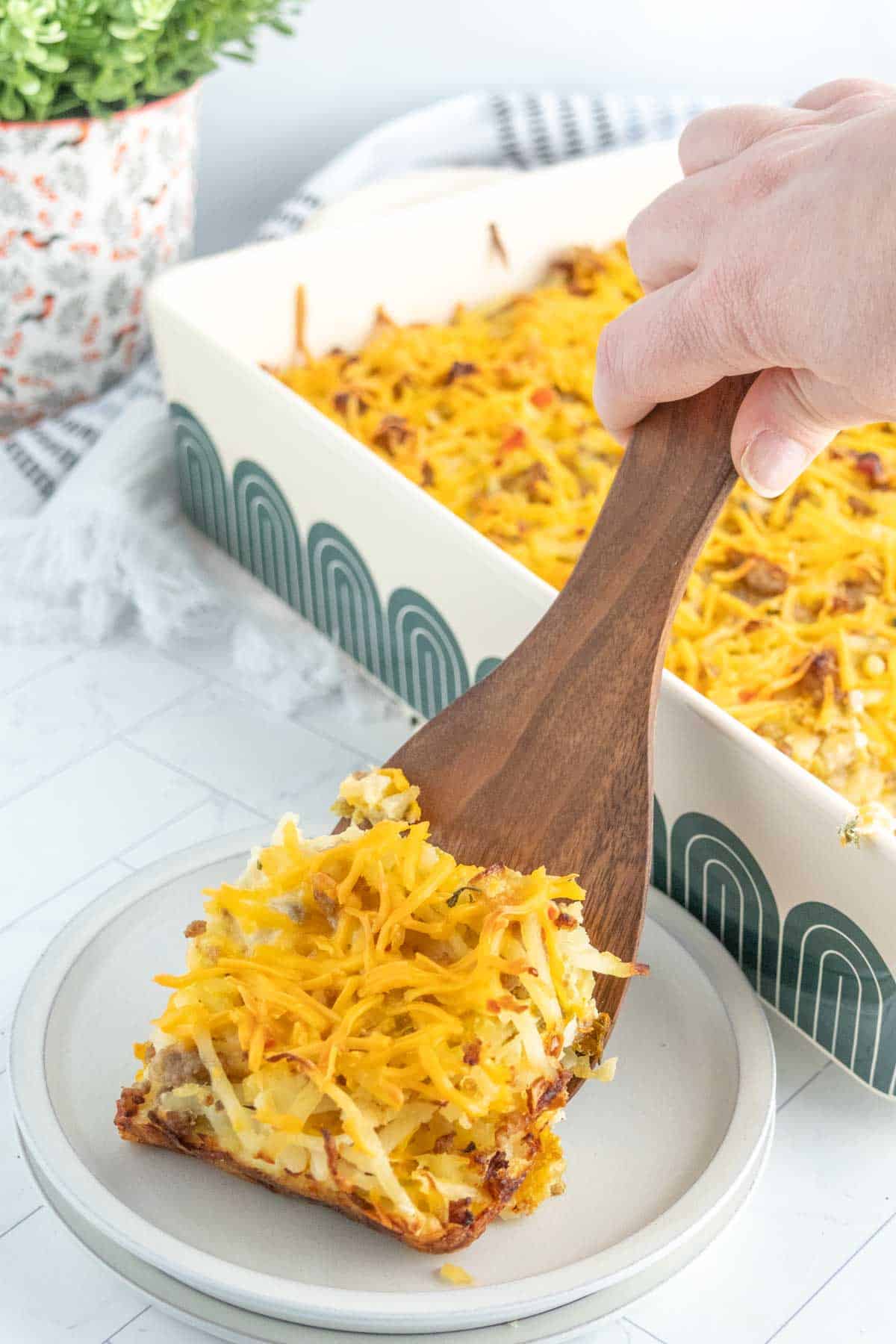 A hand uses a wooden spatula to serve a slice of cheesy breakfast casserole from a rectangular dish onto a white plate.
