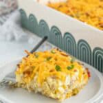 A slice of breakfast casserole topped with shredded cheese and herbs sits on a plate with a fork, while the rest of the savory breakfast casserole remains in a baking dish in the background.