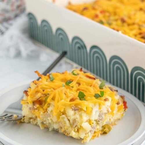 A slice of breakfast casserole topped with shredded cheese and herbs sits on a plate with a fork, while the rest of the savory breakfast casserole remains in a baking dish in the background.