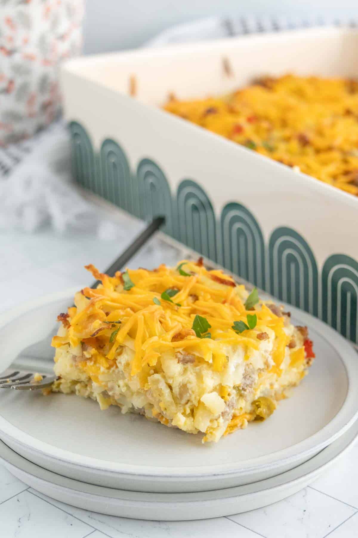 A slice of breakfast casserole topped with shredded cheese and herbs sits on a plate with a fork, while the rest of the savory breakfast casserole remains in a baking dish in the background.