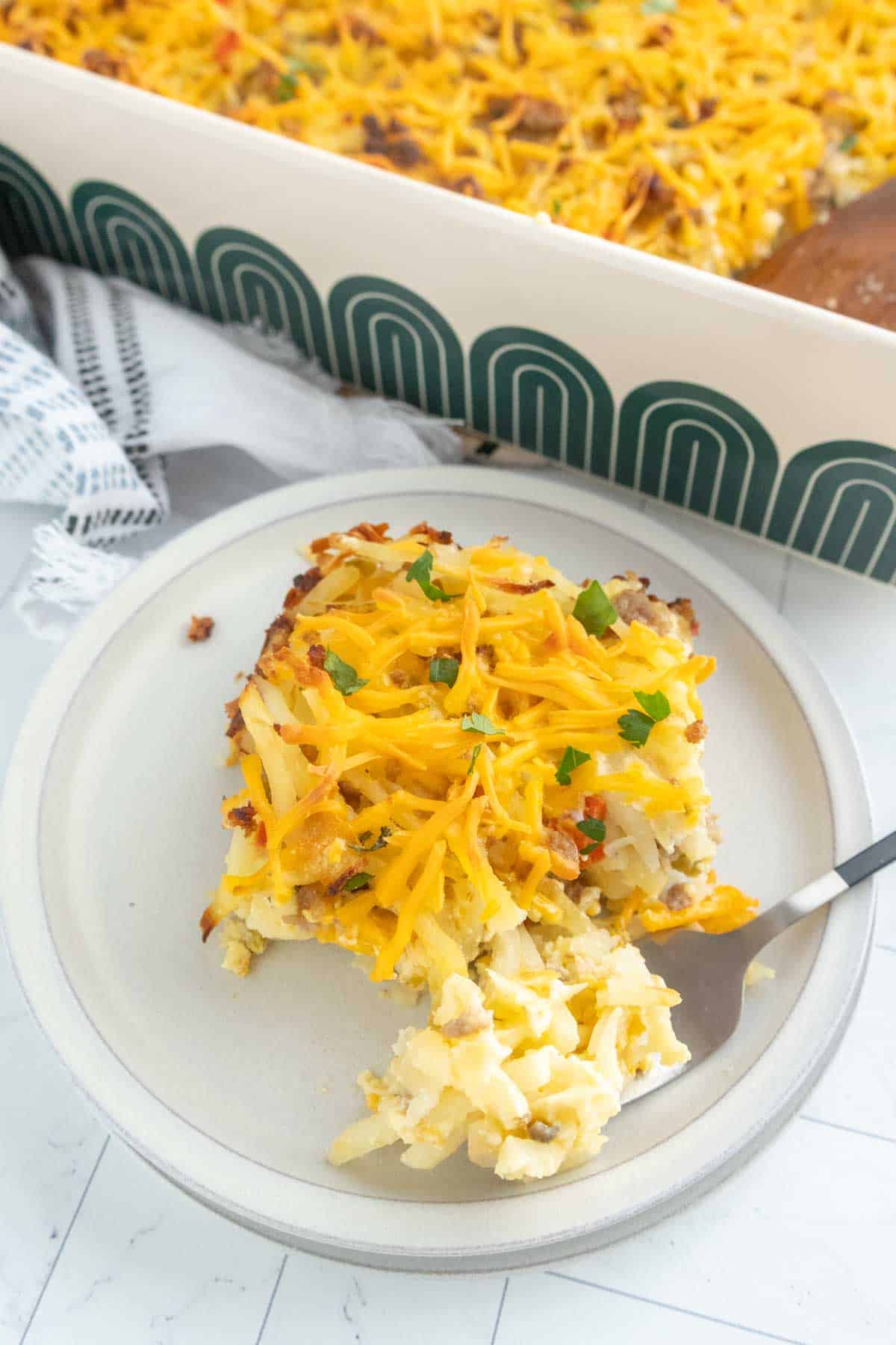 A plate with a serving of cheesy breakfast casserole, topped with shredded cheddar and green garnish, sits beside a baking dish filled with more hash brown casserole.