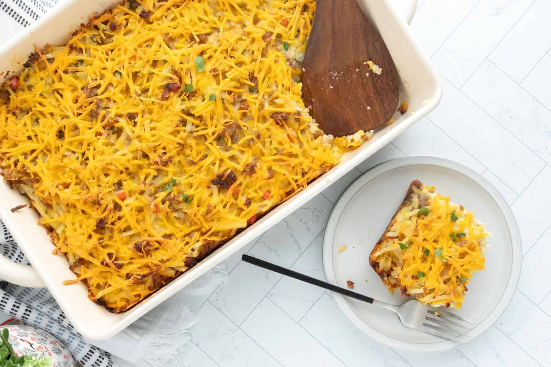 A baked breakfast casserole of hash browns topped with shredded cheese in a white dish, with a serving on a plate beside it and a fork.