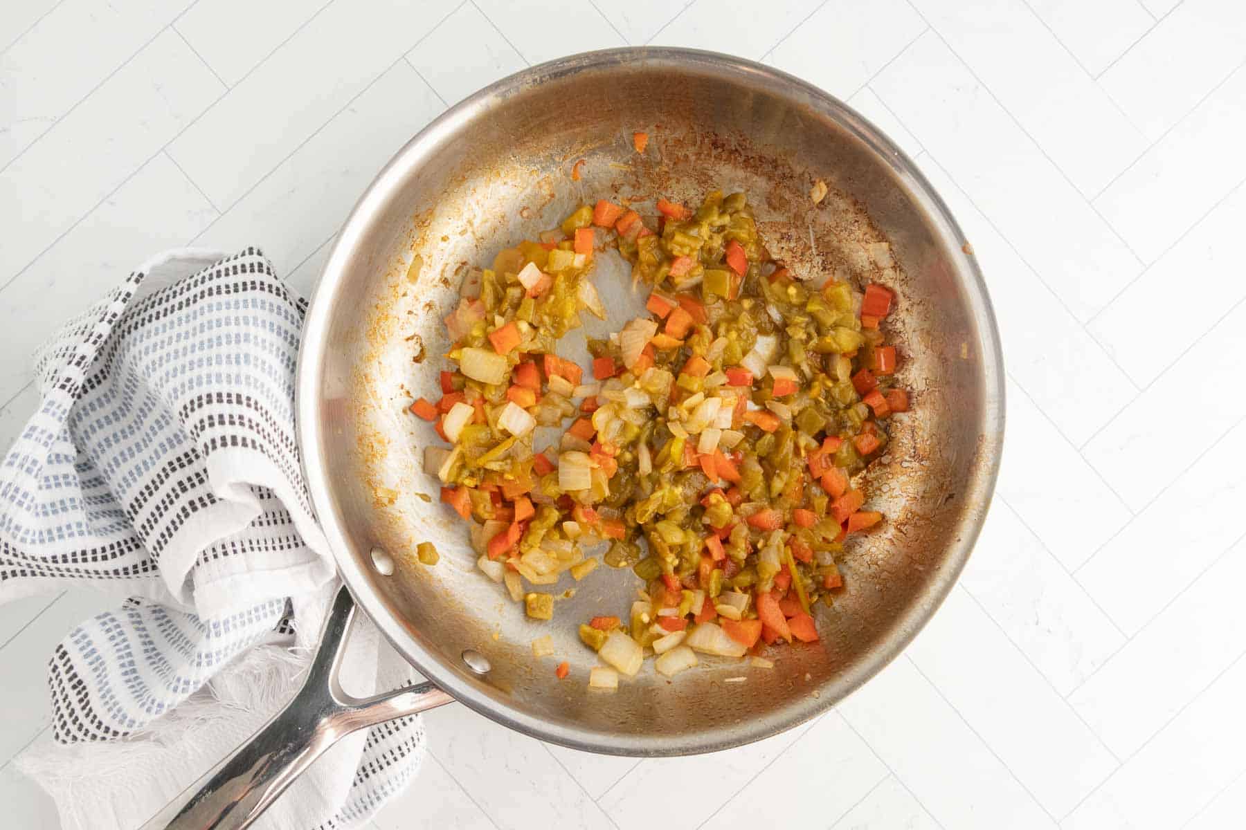 Scrambled vegetables cooking in a stainless steel skillet on a white tiled countertop.