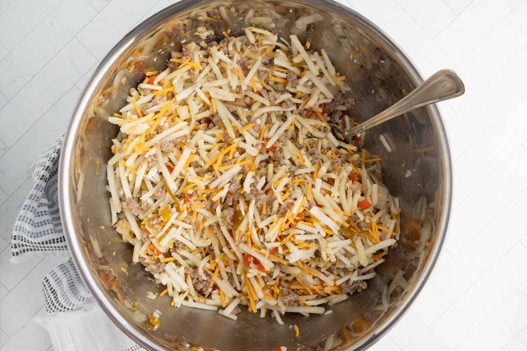 A large metal bowl filled with uncooked shredded hash browns, ground meat, diced peppers, and shredded cheese sits ready for a delicious breakfast casserole, a spoon inside for mixing all the hearty ingredients.
