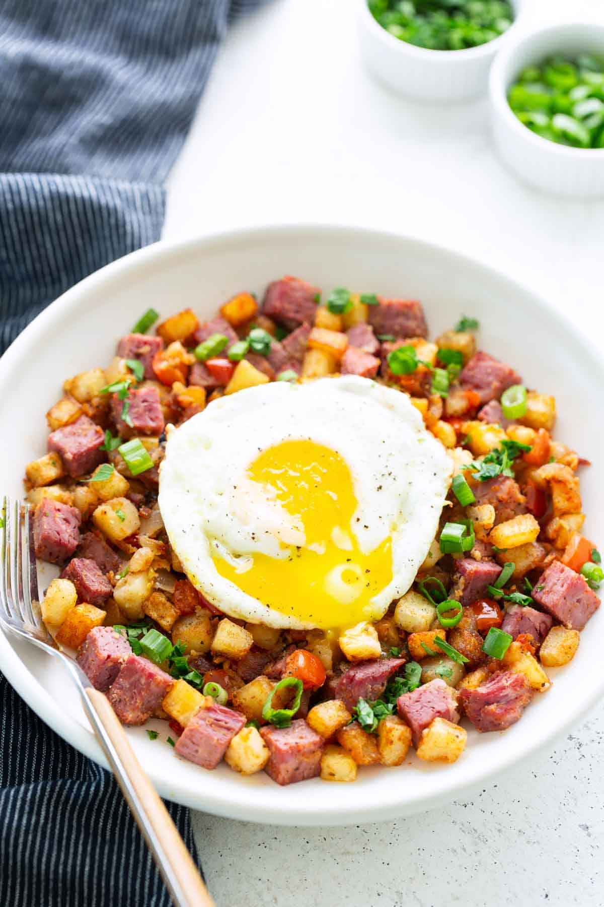 A hearty bowl of corned beef hash with diced potatoes, fresh green onions, and a sunny-side-up egg on top, served with a fork on the side.