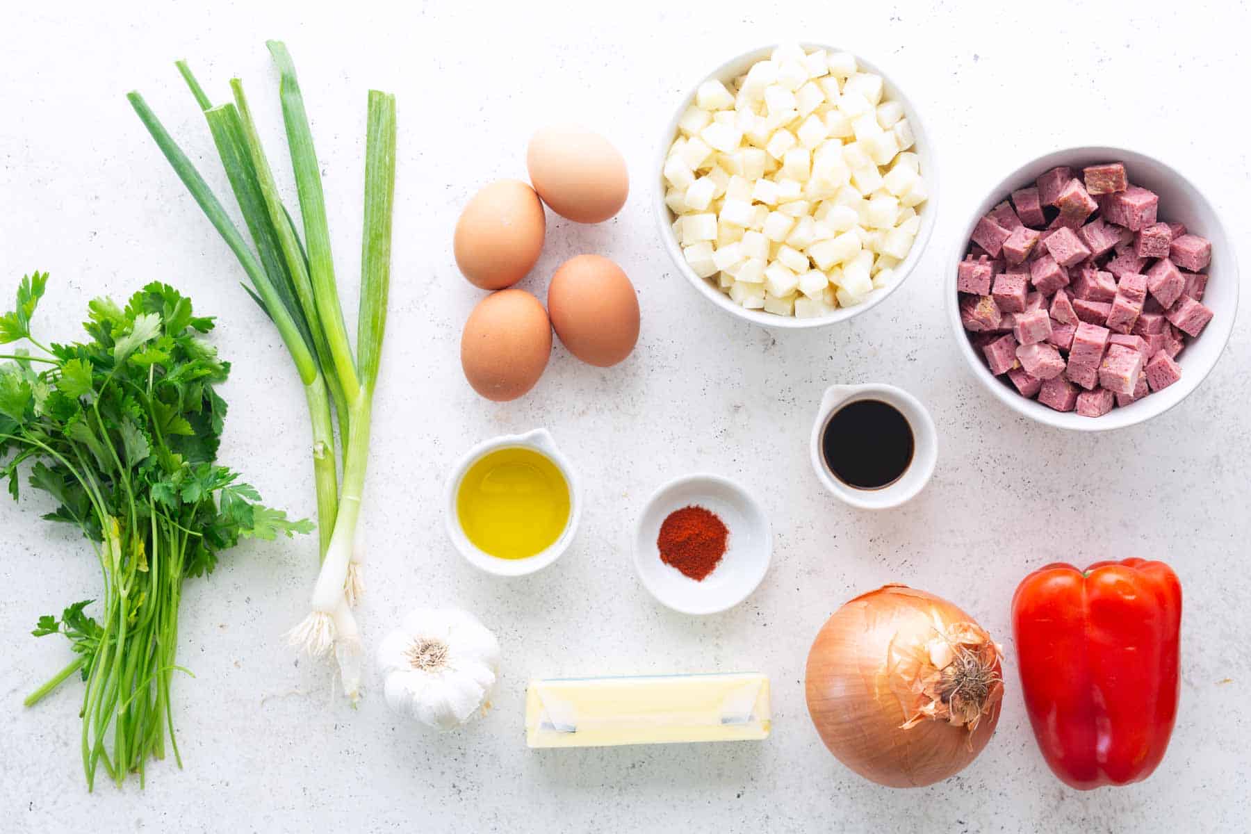 Various ingredients on a white surface, including parsley, green onions, eggs, diced potatoes, diced ham or corned beef hash, olive oil, paprika, Worcestershire sauce, garlic, butter, onion, and red bell pepper.