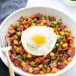 A plate of classic corned beef hash with diced potatoes and green onions, topped with a sunny-side-up egg, served with a fork on the side.