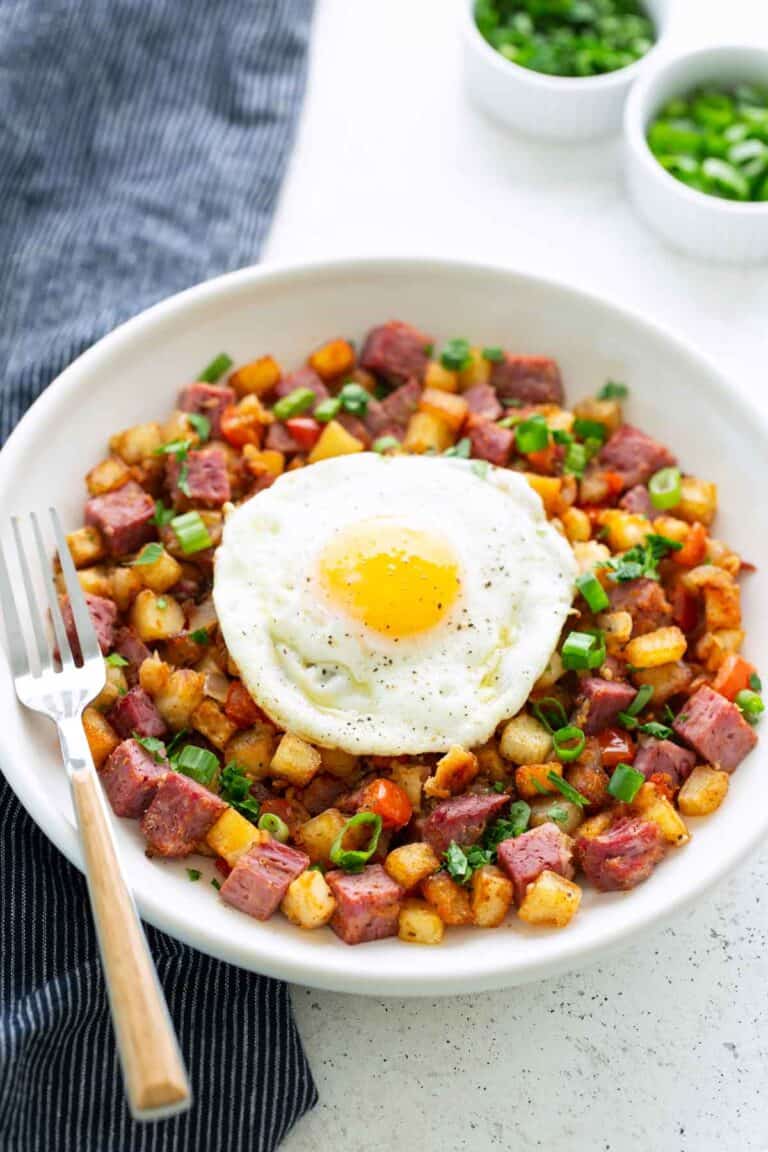 A plate of classic corned beef hash with diced potatoes and green onions, topped with a sunny-side-up egg, served with a fork on the side.