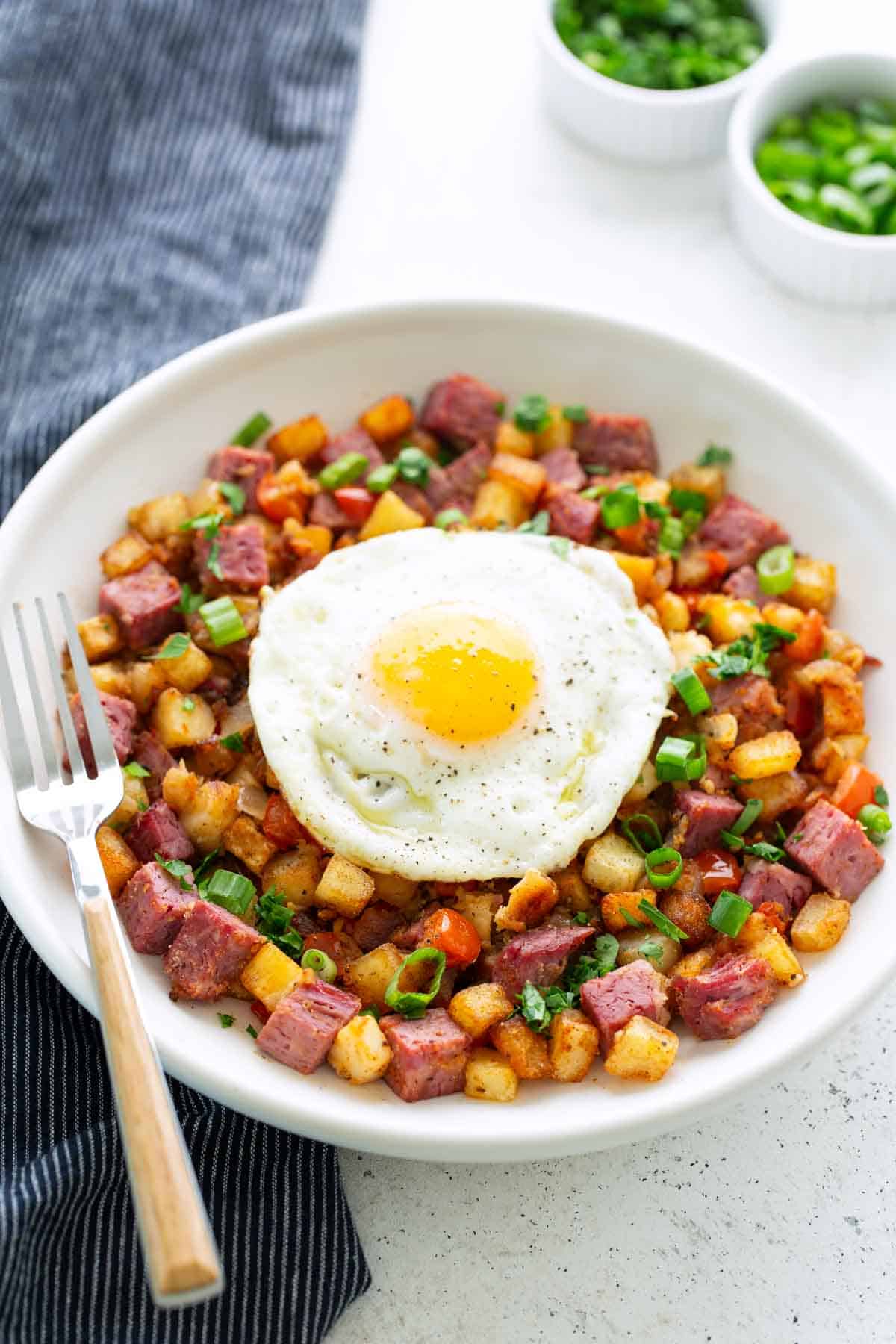 A plate of classic corned beef hash with diced potatoes and green onions, topped with a sunny-side-up egg, served with a fork on the side.