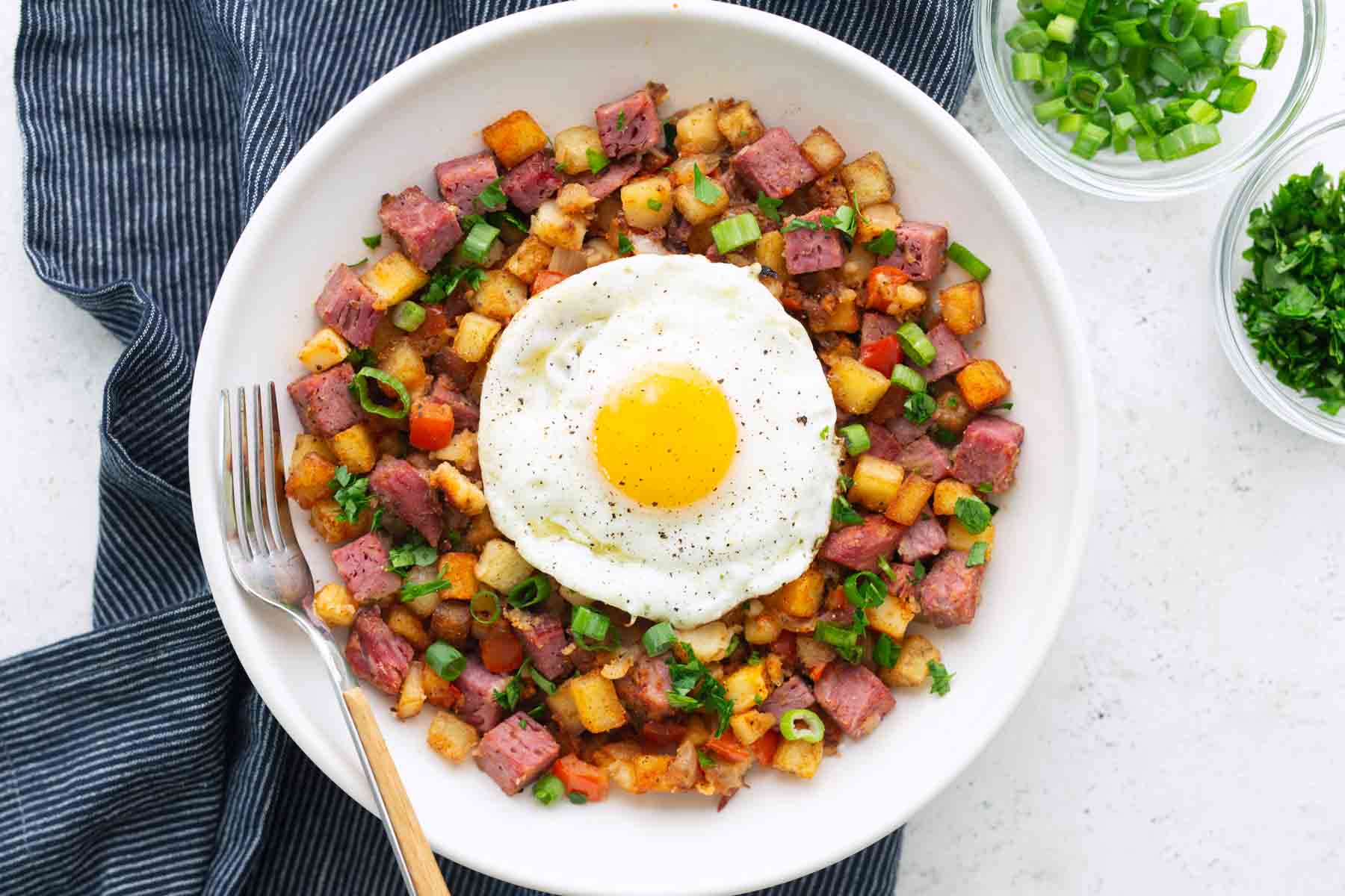 A plate of classic corned beef hash topped with a sunny-side-up egg, garnished with chopped green onions, with a fork and bowls of fresh herbs on the side.