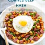 A plate of corned beef hash with diced potatoes and green onions, topped with a sunny-side-up egg, next to a fork and a small bowl of chopped green onions.
