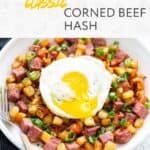 A plate of corned beef hash with diced potatoes, bell peppers, and green onions, topped with a sunny side up egg. A fork rests on the side of the plate.