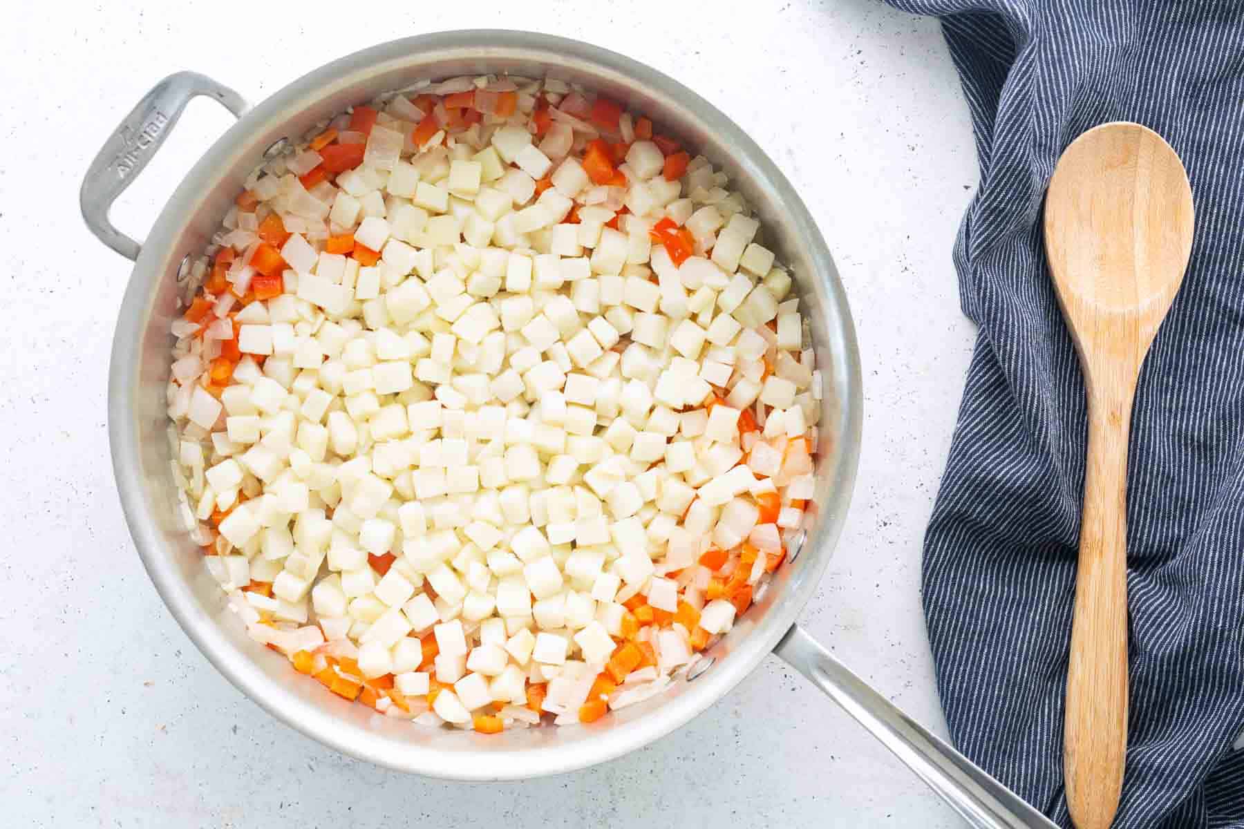 A saucepan filled with diced potatoes and carrots for corned beef hash sits on a white surface next to a blue-striped cloth and a wooden spoon.