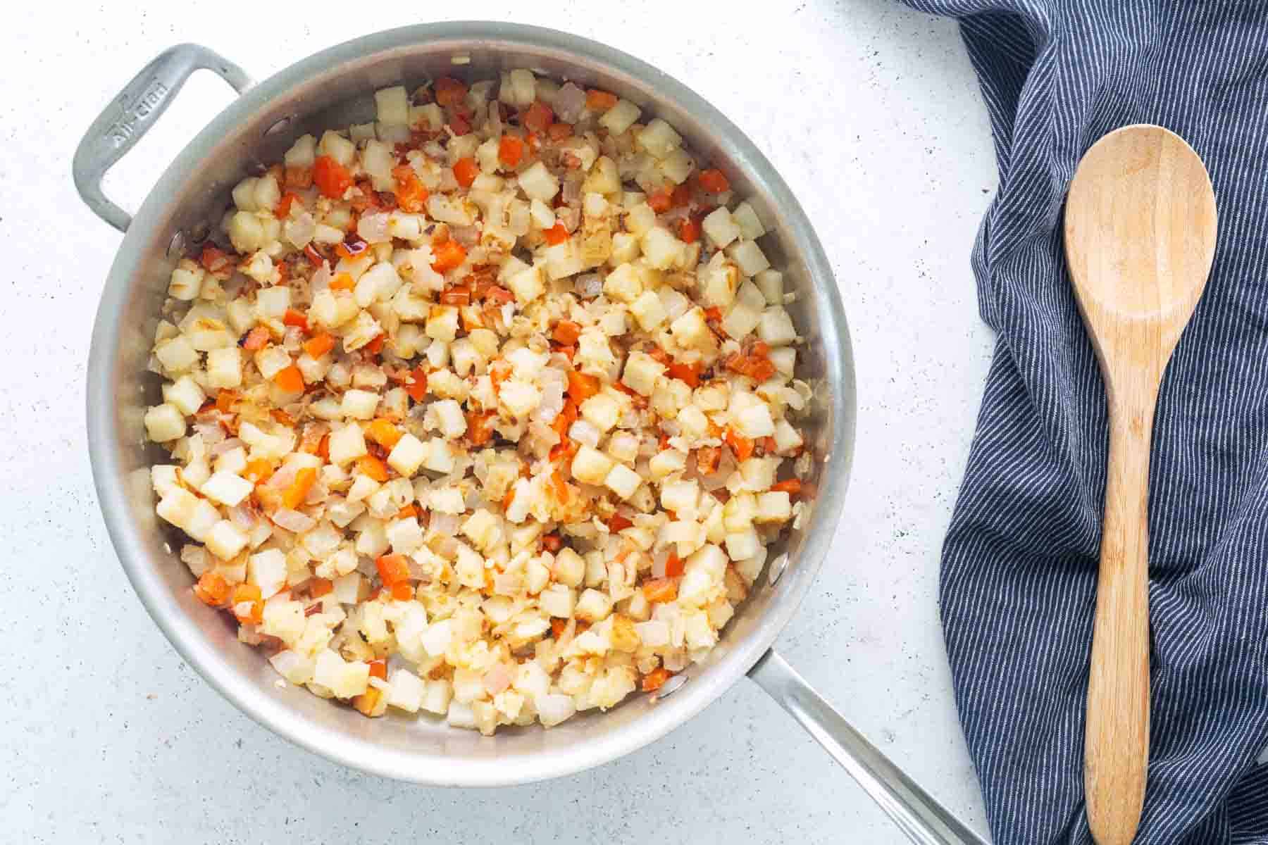 A sauté pan filled with diced potatoes, carrots, onions, and savory corned beef hash sits on a white countertop next to a wooden spoon and a blue-striped cloth.