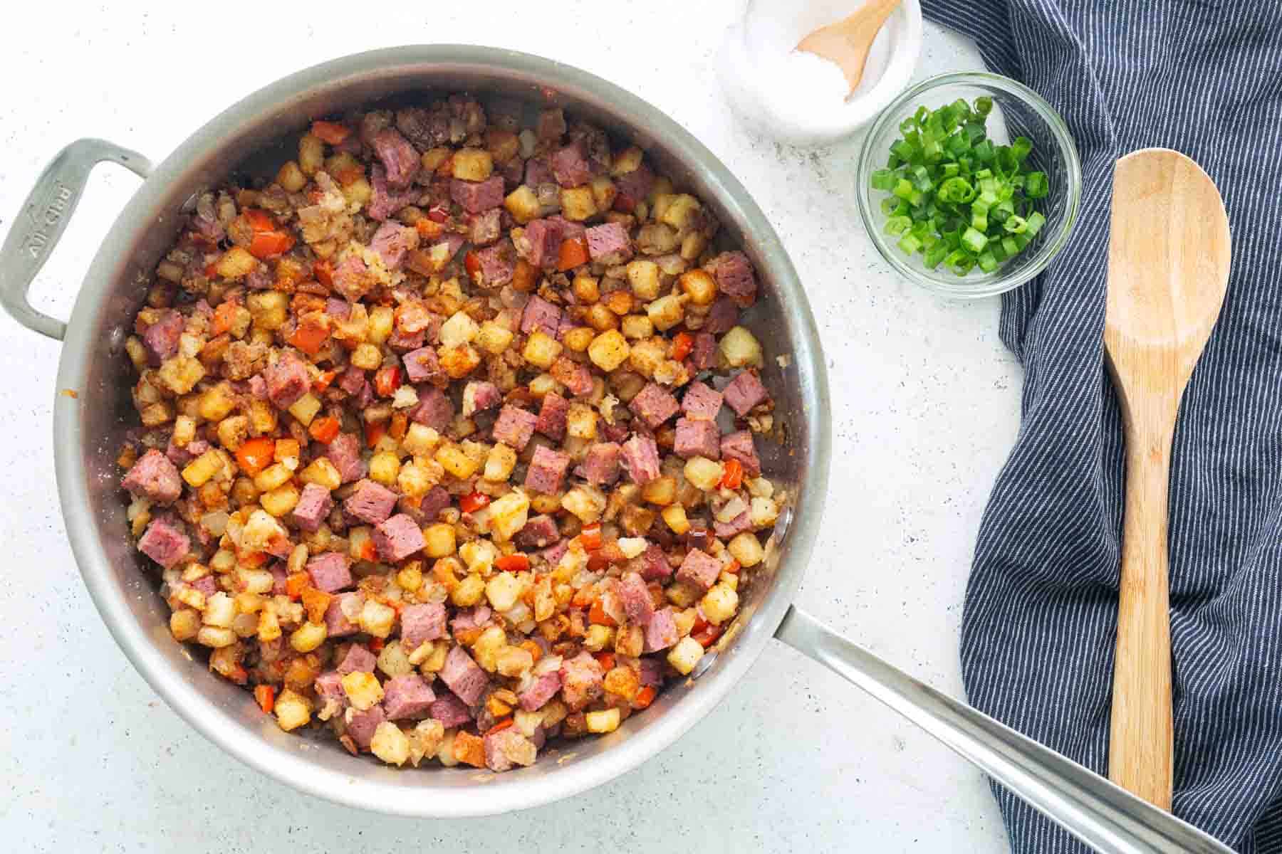 A skillet filled with classic corned beef hash—diced corned beef, potatoes, and vegetables—sits on a countertop next to chopped green onions, salt, a wooden spoon, and a striped kitchen towel.