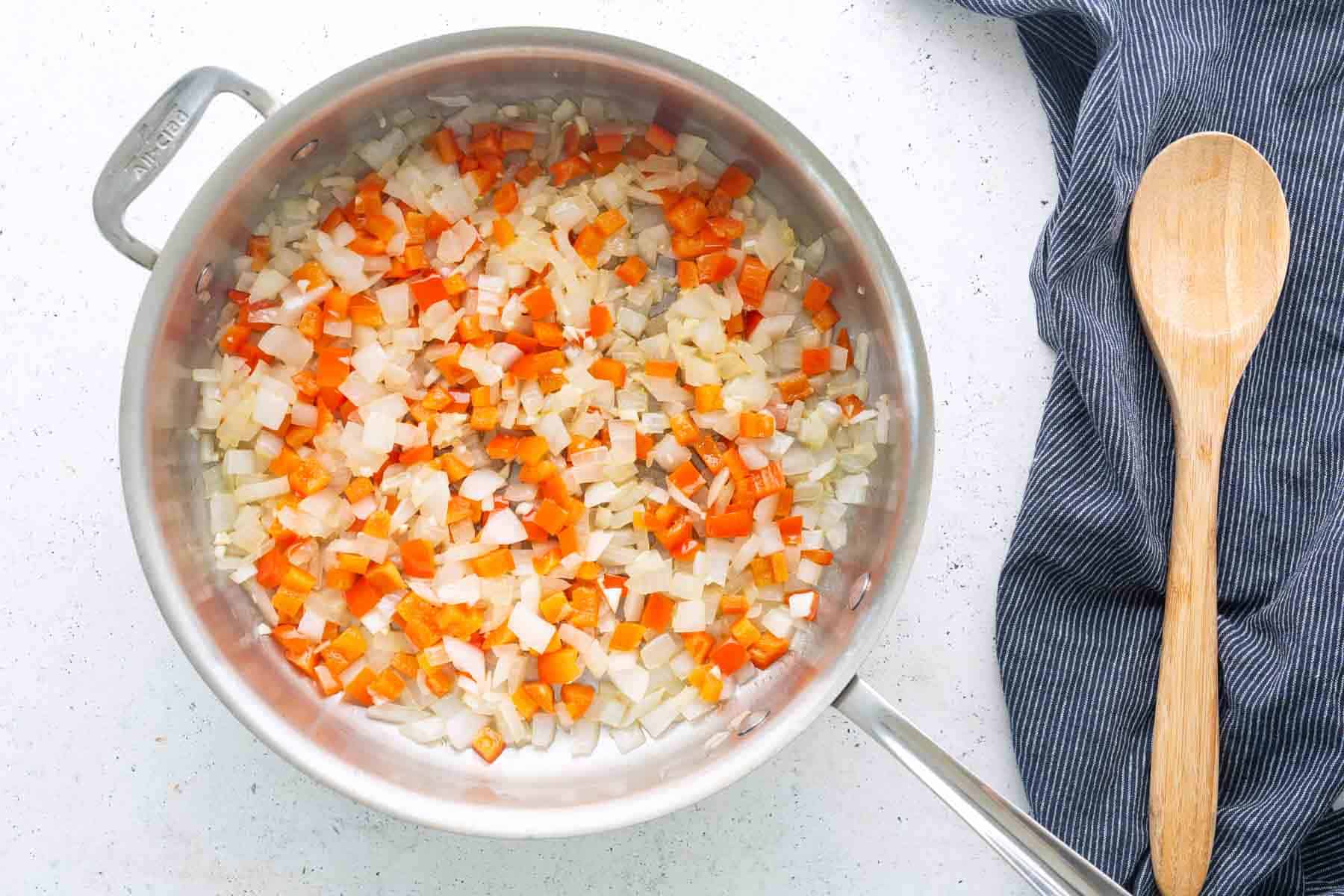 Chopped onions and red bell peppers sautéing in a stainless steel pan, the perfect start for a savory corned beef hash, with a wooden spoon resting on a striped cloth.