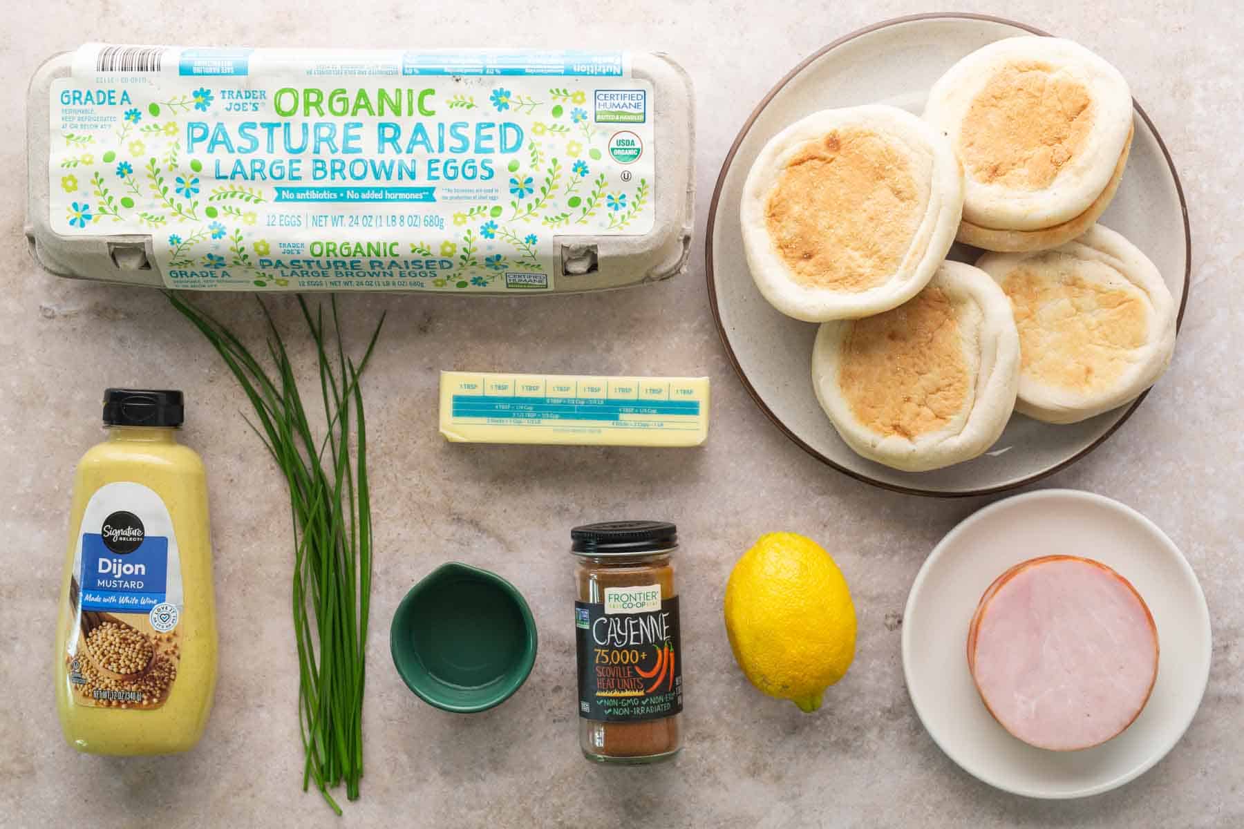 Eggs Benedict ingredients—including eggs, English muffins, ham, Dijon mustard, butter, cayenne pepper, lemon, and chives—are artfully arranged on a light surface.