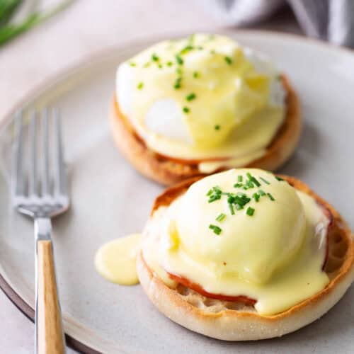 Two classic eggs benedict on English muffins topped with silky hollandaise sauce and chopped chives, served on a plate with a fork.