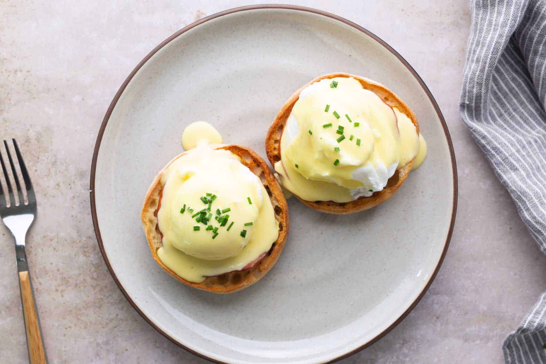 Two classic eggs Benedict on toasted English muffins with rich hollandaise sauce and chopped chives, served on a ceramic plate with a fork beside it.