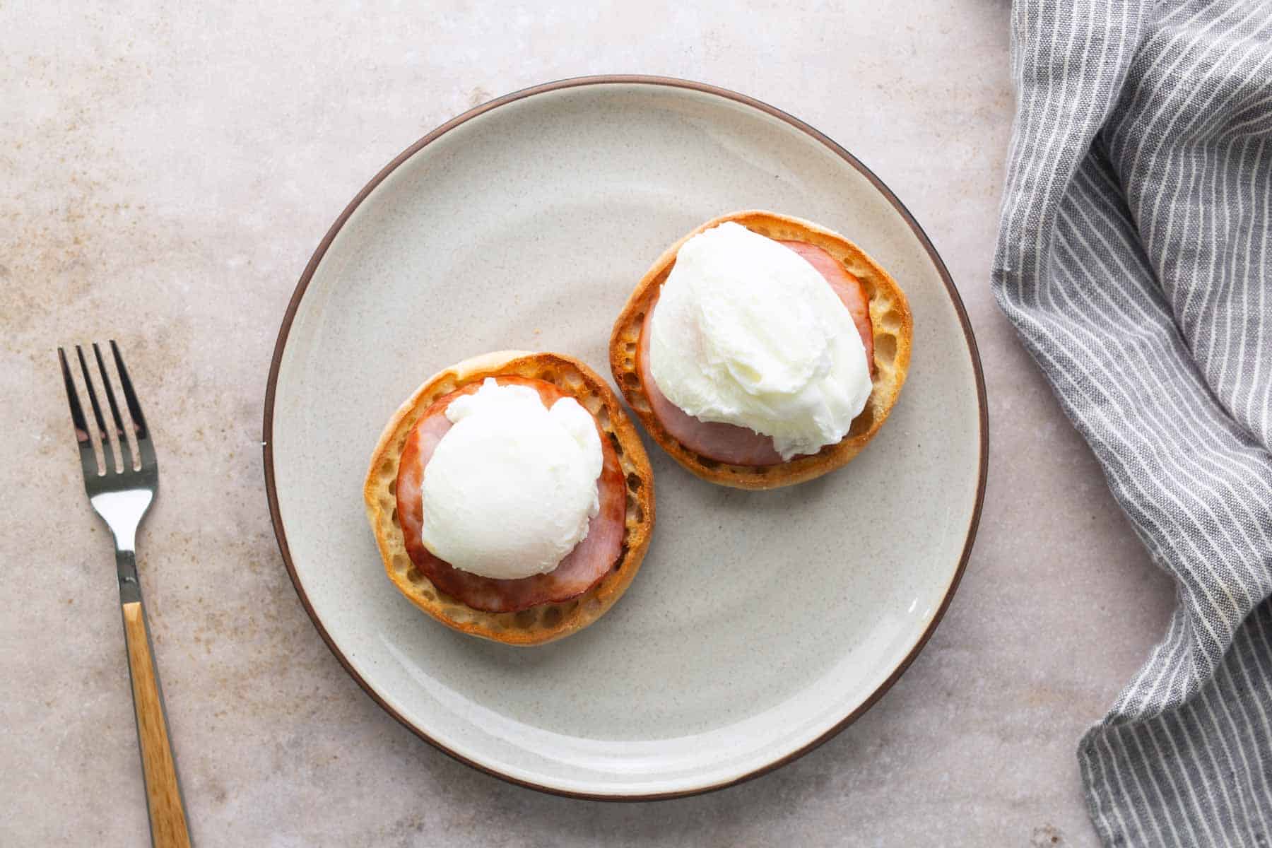 A plate with two English muffins topped with ham and poached eggs, creating a classic eggs benedict, sits next to a fork and a striped cloth napkin.